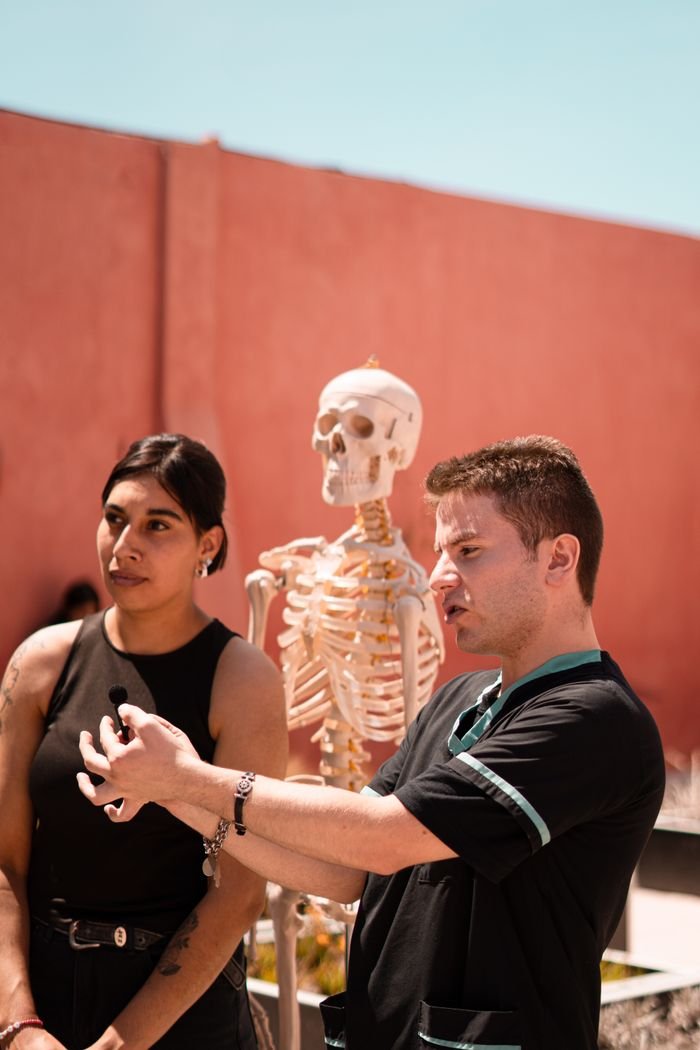 hero-gallery-03 Two medical students study a human skeleton outdoors, explaining anatomy concepts under bright sunlight.